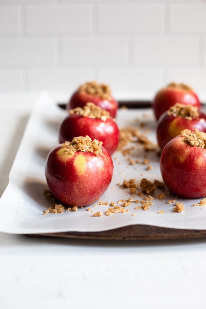 A baking sheet is covered with parchment and bright red apples that have been stuffed with a filling of oats, flour, and brown sugar.