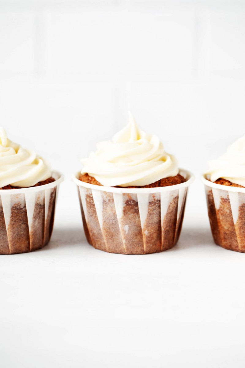 Three vegan cupcakes, made with carrots and cream cheese frosting, are lined up neatly in front of a white brick surface.