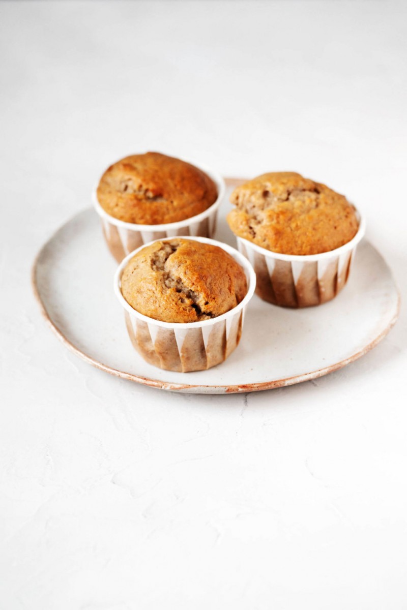 A small, white ceramic plate with a tan rim holds three freshly baked muffins in their muffin liners.