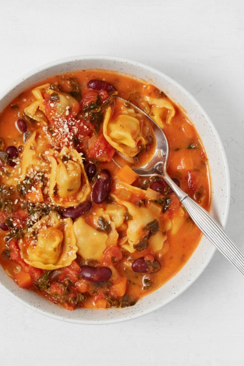 Vegan tortellini soup has been placed into a white bowl. It's being served with a silver spoon.