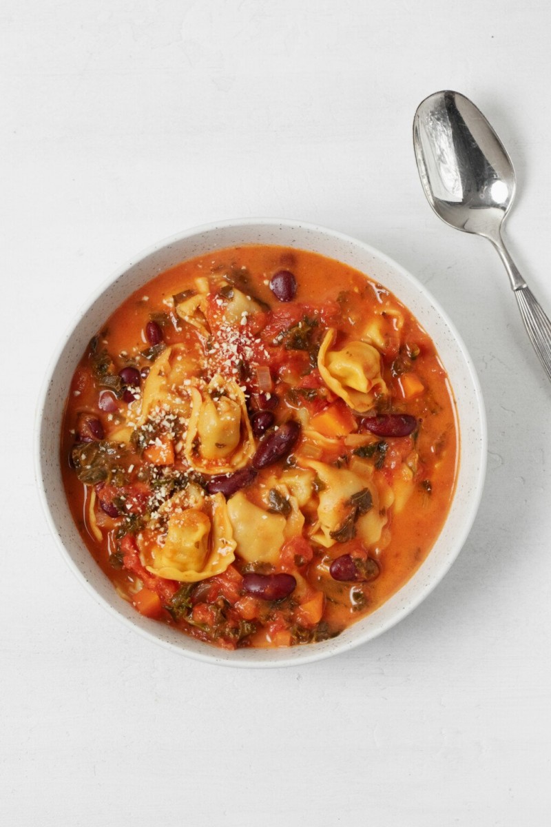 A round, white bowl of vegan tortellini soup has been topped with plant-based parmesan cheese. It rests on a white surface with a silver spoon nearby.