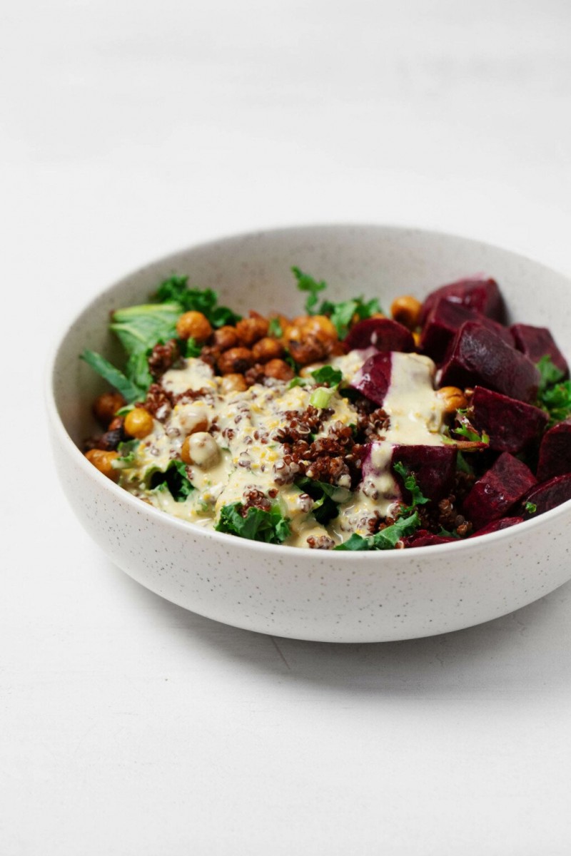 An angled photograph of a round, white ceramic bowl, which is filled with beets, quinoa, and crispy chickpeas, along with a creamy white dressing.