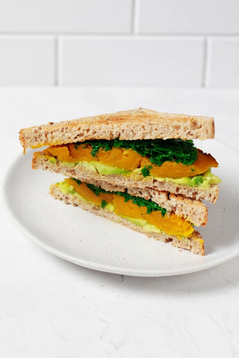 An angled image of a vegan sandwich made with layers of roasted kabocha squash, avocado, and kale. The sandwich is served on a small, round white plate.