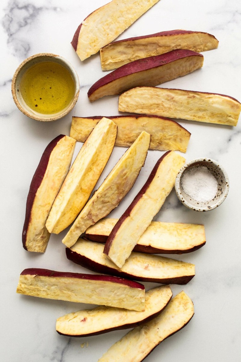 Long cut spears of Japanese sweet potato are arranged on a marble surface. A small salt cellar and pinch pot of oil are nearby.