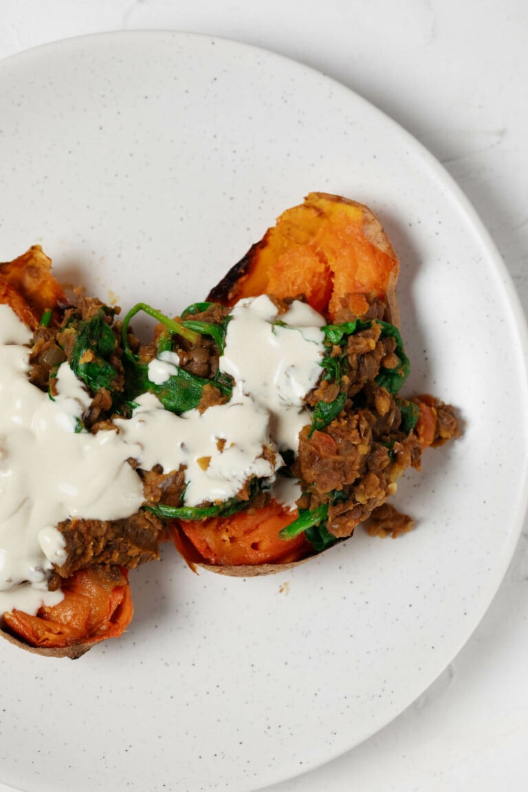 Two halves of sweet potatoes are topped with spiced lentils and a creamy tahini dressing. The potatoes rest on a round white plate.