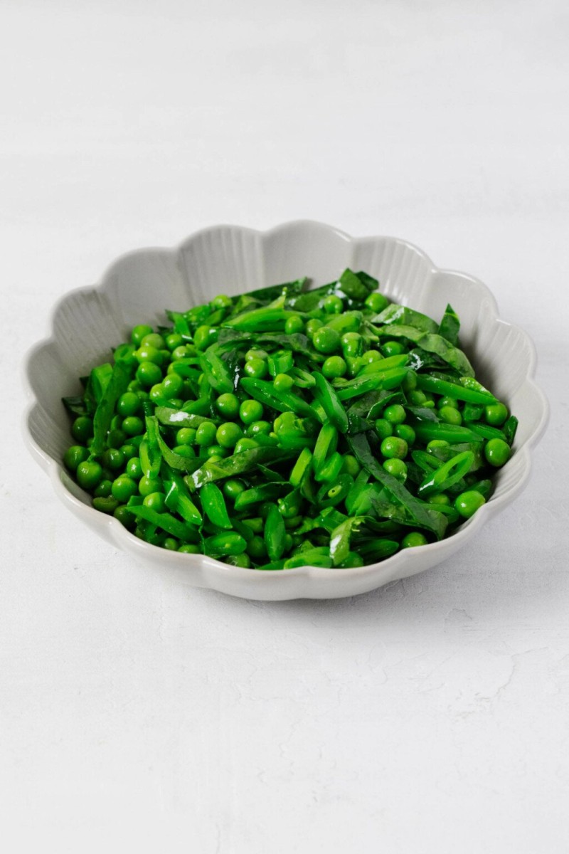 A round, fluted white bowl is filled with bright green, fresh peas and thinly sliced snap peas, along with spinach. The vegetables glisten lightly with a vinaigrette.